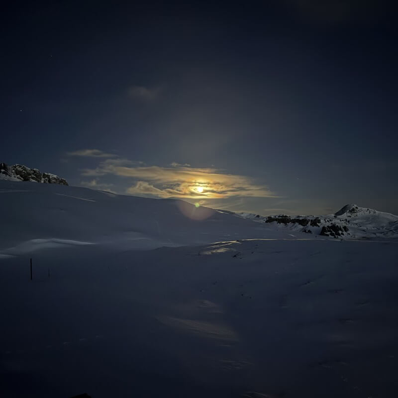 Heller Mond hinter Wolken über einer verschneiten alpinen Hochebene.