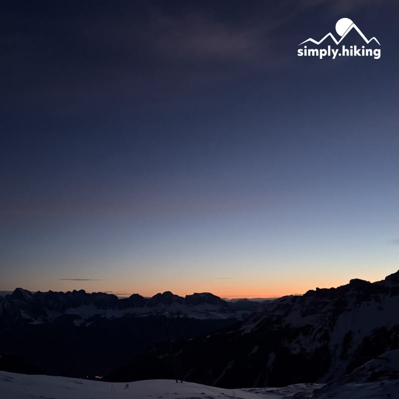 Orangefarbener Horizont hinter dunklen Bergsilhouetten, darüber blauer Himmel und das Logo von simply.hiking.