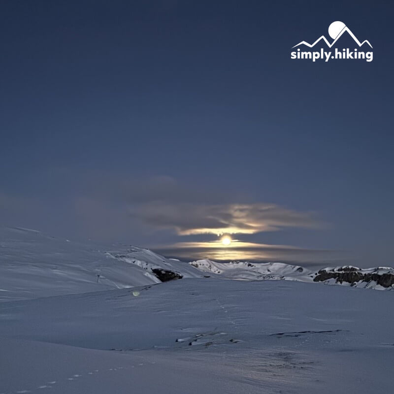 Heller Mond zwischen Wolken über verschneiten Hügeln, oben rechts das Logo von simply.hiking.