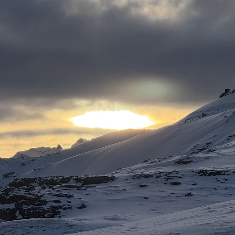 Goldenes Sonnenlicht unter dunklen Wolken über verschneiten Berghängen und Felsen.